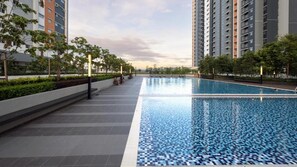 Una piscina con borde de azulejos, rodeada por un paseo y vegetación, con edificios altos al fondo.