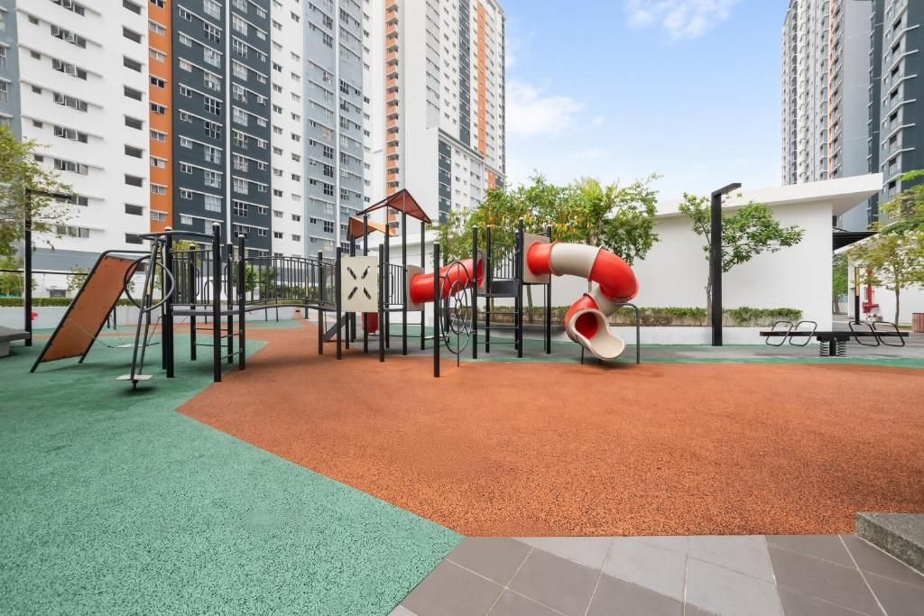 A playground with a slide and climbing structures, surrounded by a green carpet and a red rubberized area, with high-rise buildings in the background.