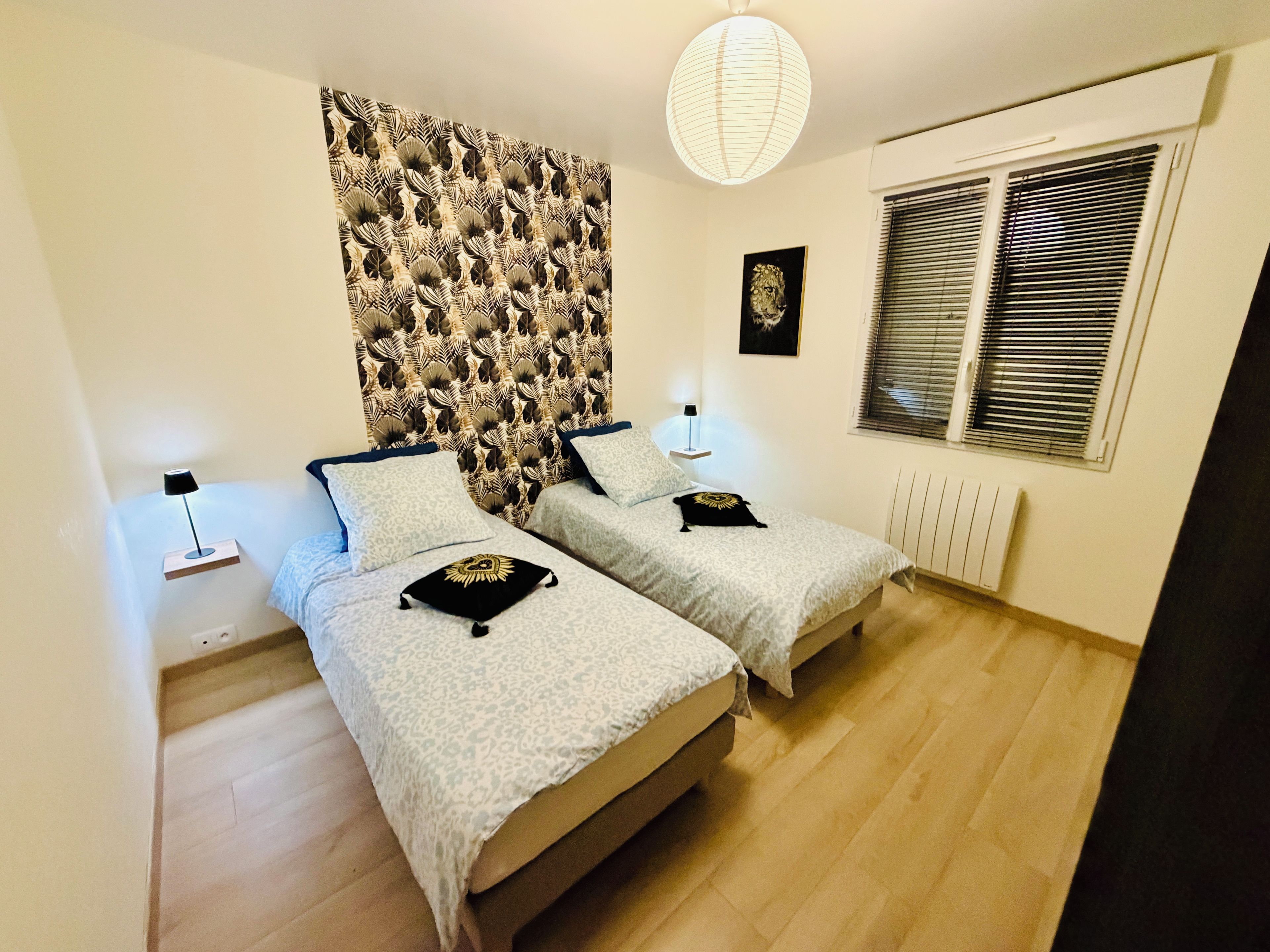 Et soveværelse med to senge, et mønstret sengegavl, sengeborde med lamper, en radiator og et vindue med persienner.
