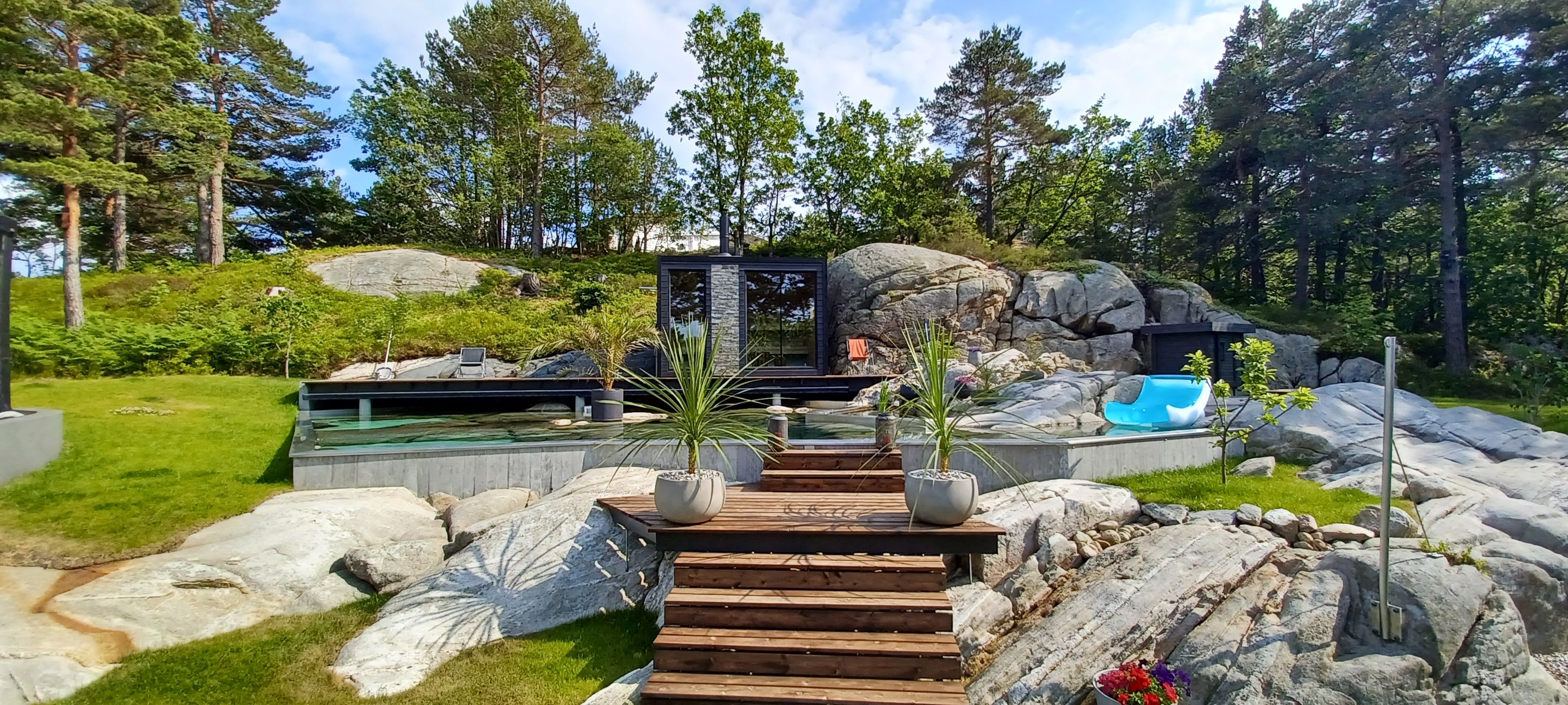 A modern outdoor pool area with a wooden deck leading to a pool, surrounded by large rocks and greenery.