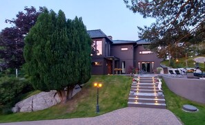 Una moderna casa a due piani con vialetto lastricato che conduce a un ingresso, circondata da un prato curato e alberi imponenti.