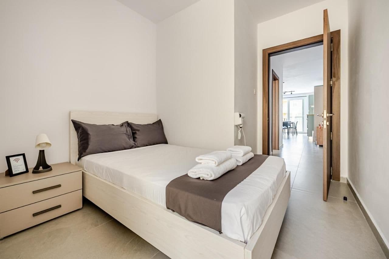 Habitación de hotel con cama, mesita de noche, lámpara y vistas al pasillo.