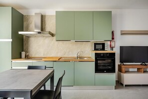 Een compacte keuken met groene kastjes, een magnetron, een oven en een eethoek met een tafel en stoelen.