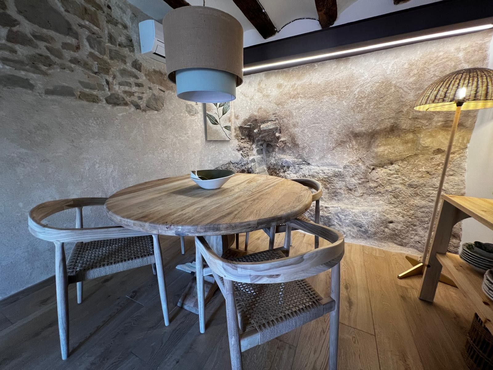 A rustic dining area with a round wooden table and wicker chairs, a hanging light fixture, and a stone wall backdrop.