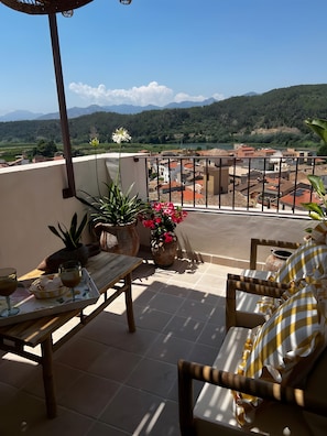 Una terraza con mesa, sillas, plantas en macetas y vistas a un pueblo y montañas.