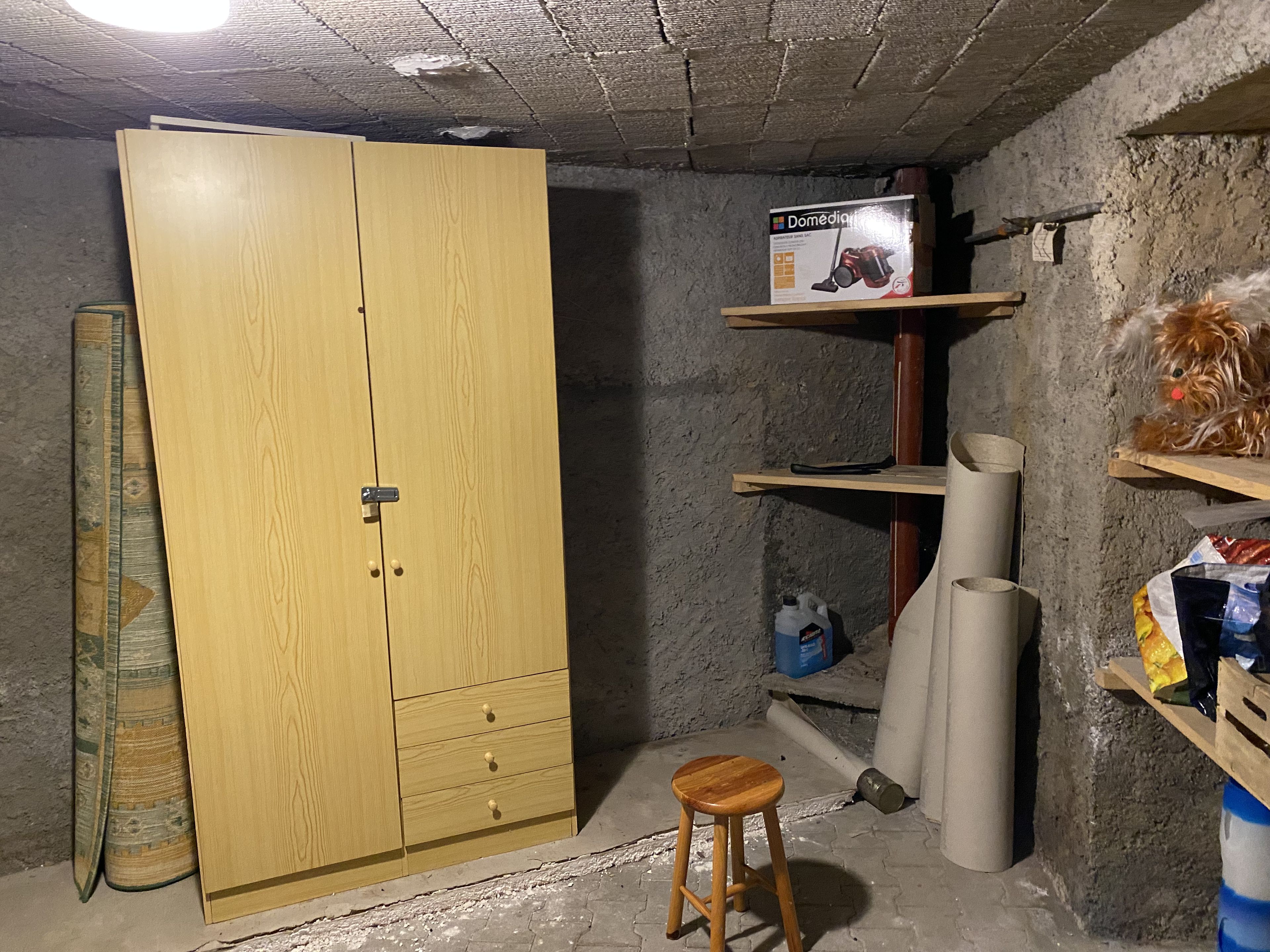 Een kamer met een houten garderobe, een klein krukje, een plank met een camera, en verschillende spullen aan de muur.