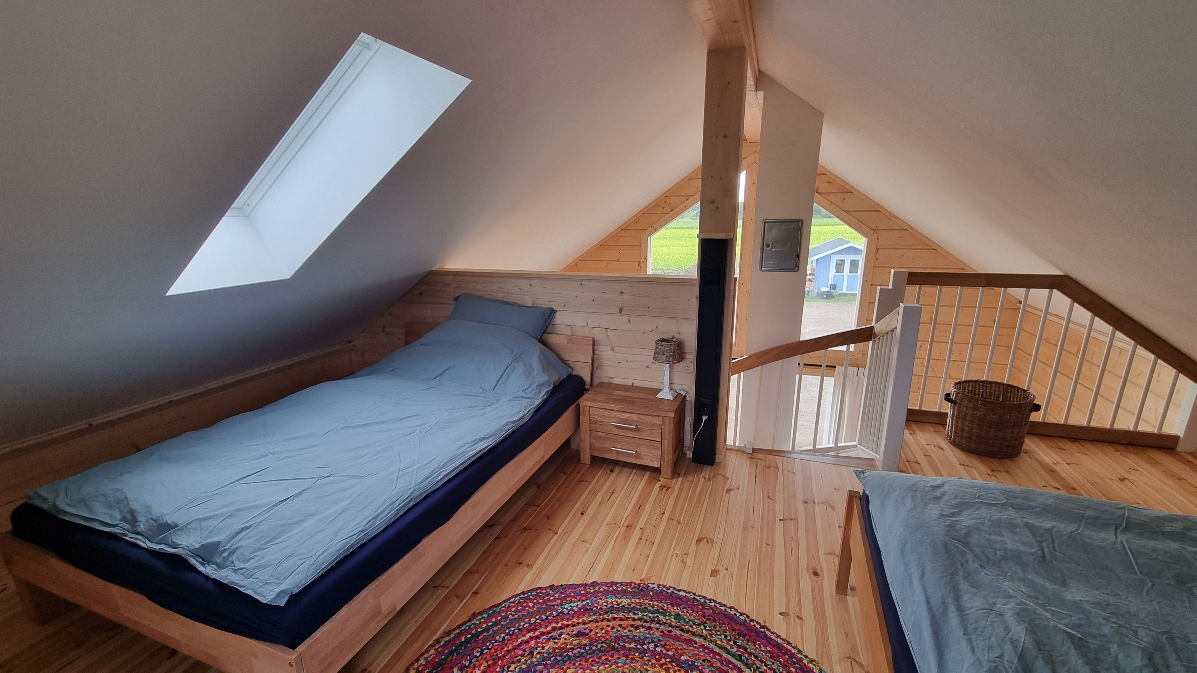 Quarto com teto inclinado, cama, mesa de cabeceira, abajur, tapete colorido e vista para o exterior.