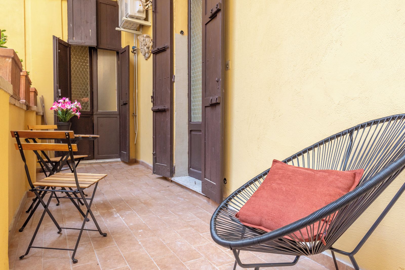 A narrow outdoor corridor with a chair and a table, a potted plant, and a door with a window.