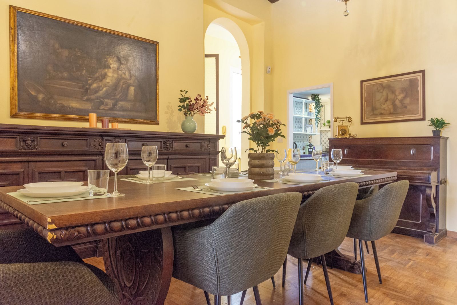A dining room with a long table set for six, flanked by chairs, a large painting above the table, and a vase with flowers on the table.