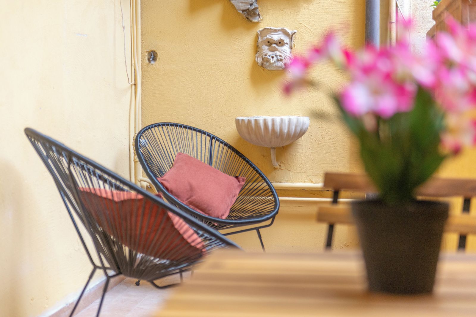 Una stanza con un tavolo di legno, due sedie in filo di ferro, un vaso con una pianta e un oggetto decorativo appeso al muro.