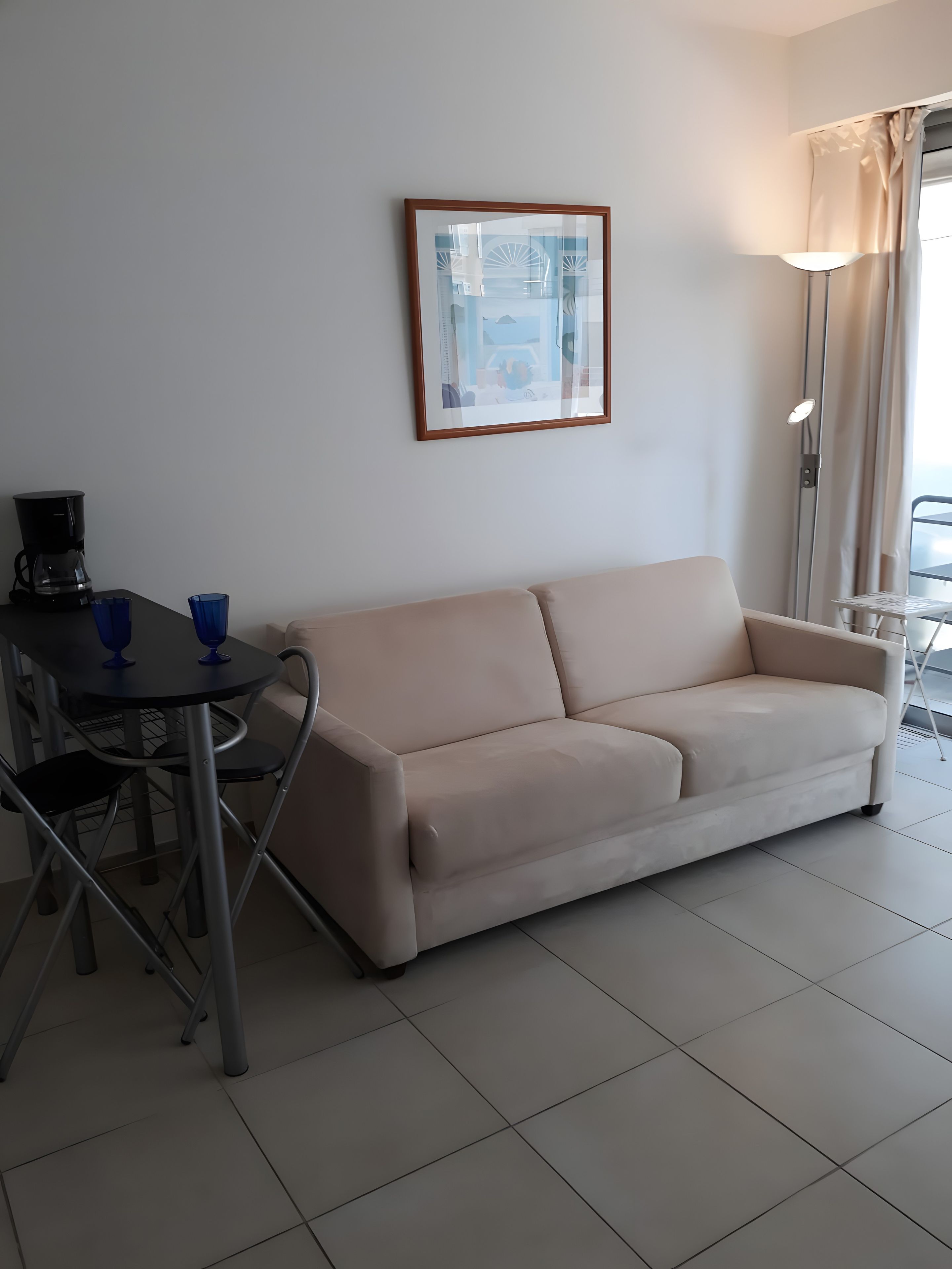 Un soggiorno con un divano beige, un tavolino rotondo con due tazze blu, una macchina da caffè nera e un quadro appeso al muro.