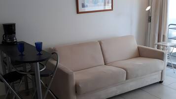 Un soggiorno con un divano beige, un tavolino rotondo con due tazze blu, una macchina da caffè nera e un quadro appeso al muro.