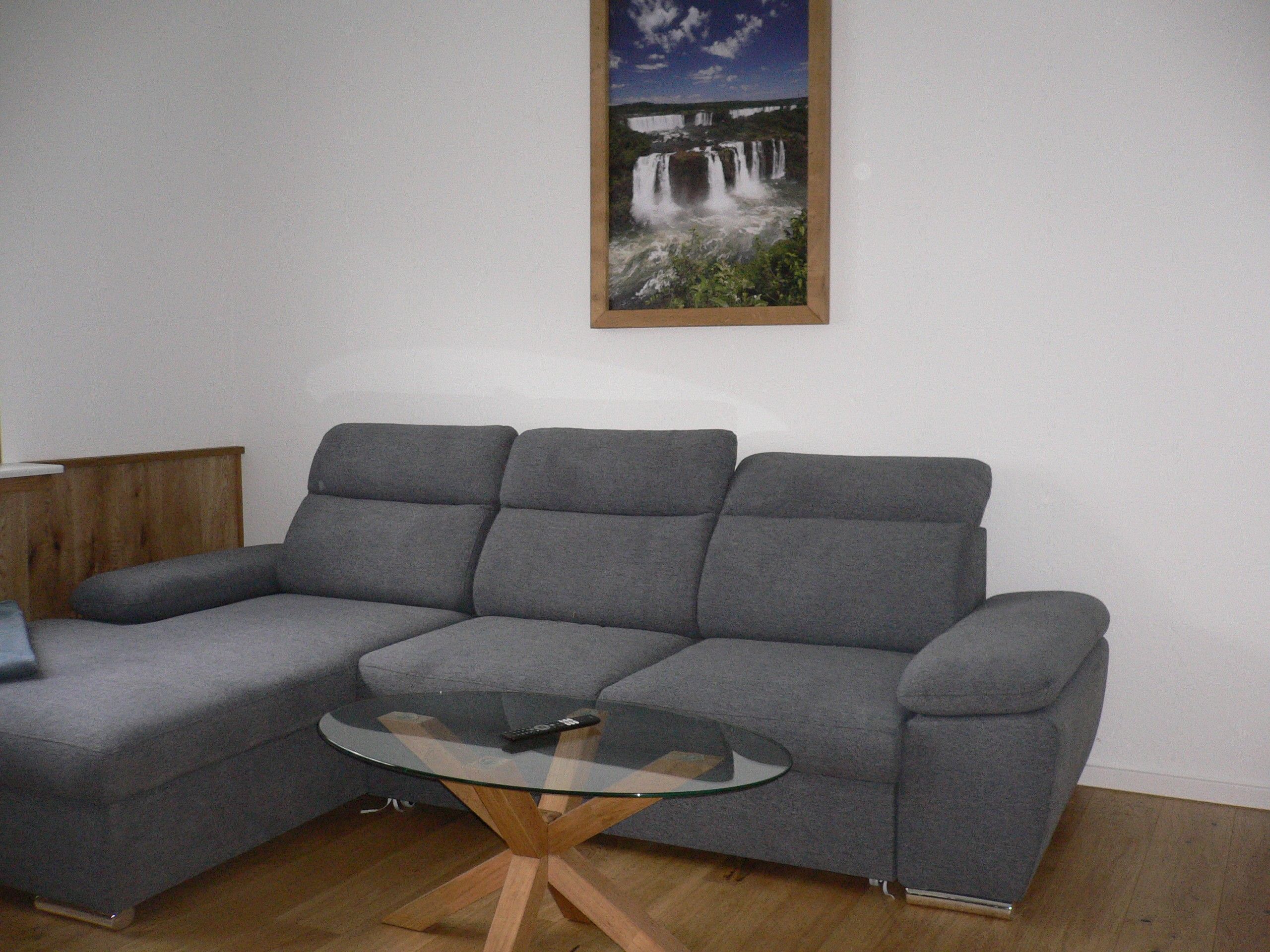 Ein graues Ecksofa mit einem Glastisch davor, einem Holztisch daneben und einem gerahmten Bild eines Wasserfalls an der Wand.