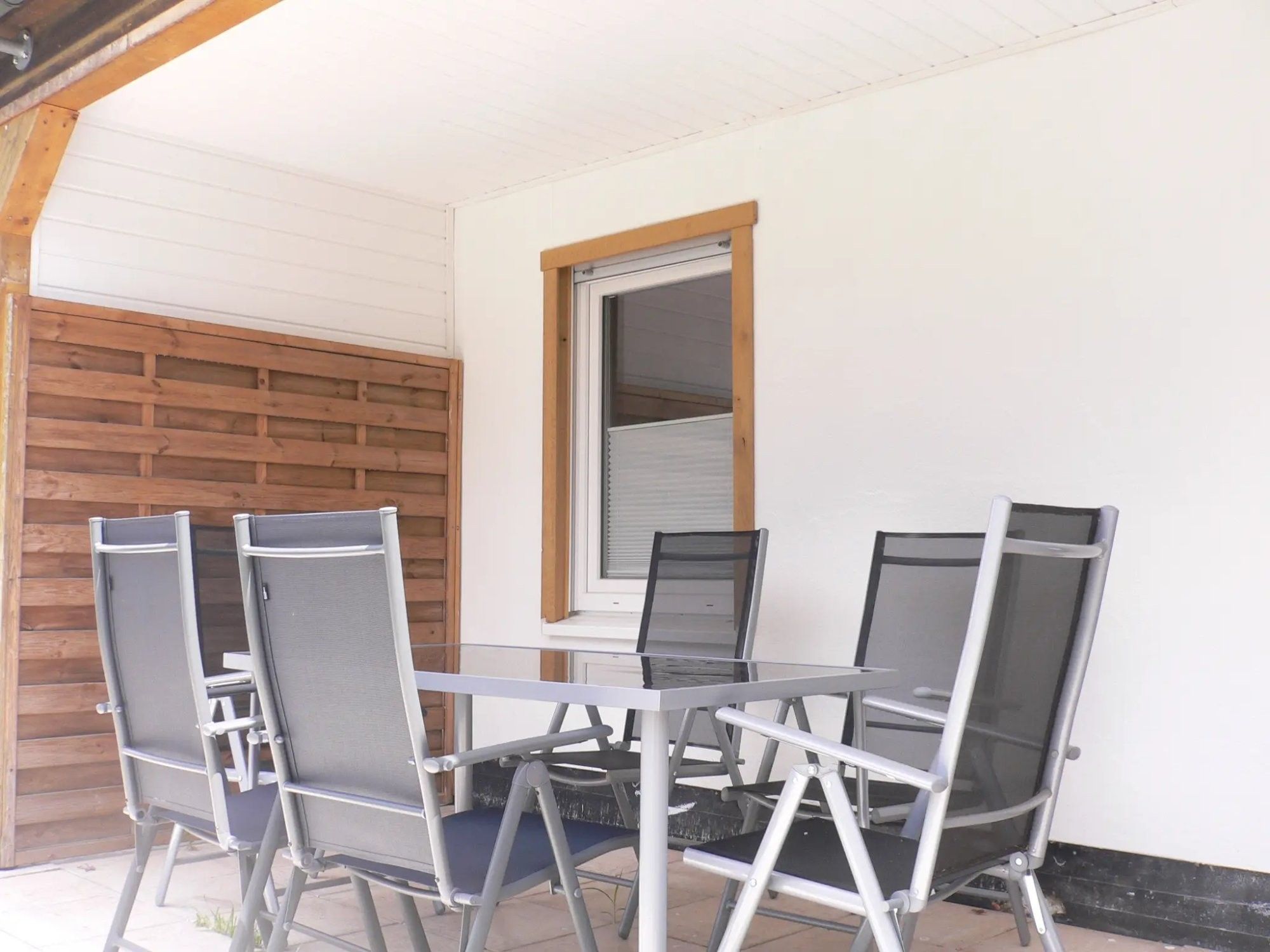Eine Terrasse mit einem Glastisch und vier Stühlen, einem Holzzaun und einem Fenster mit Jalousien.