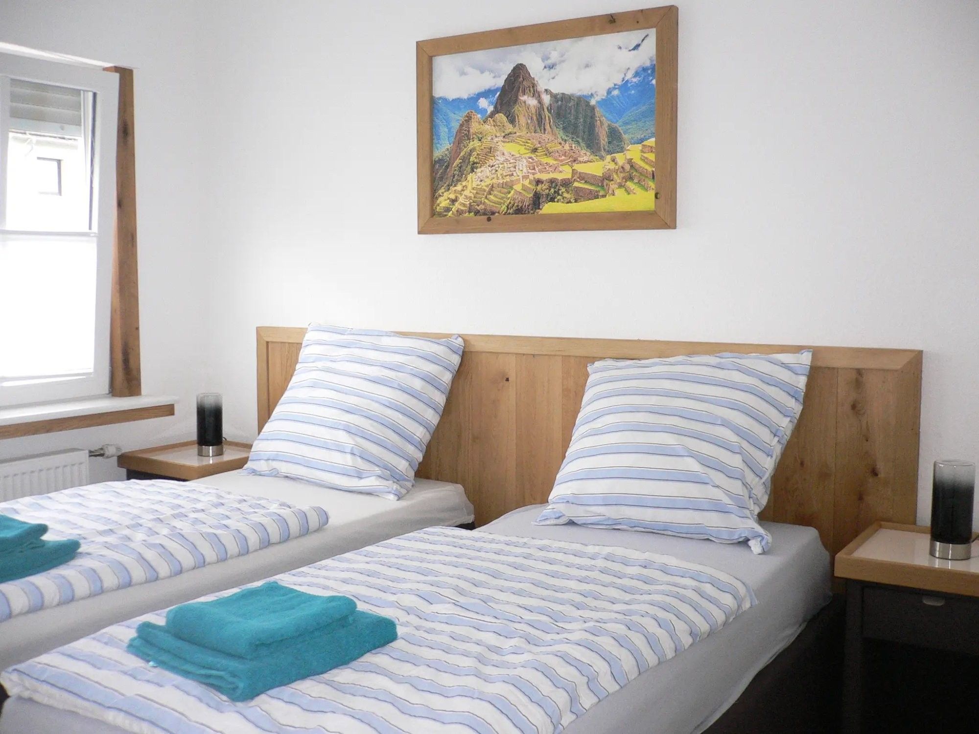 Ein Hotelzimmer mit zwei Betten, jeweils mit gestreifter Bettwäsche und einem blauen Handtuch. An der Wand hängt ein gerahmtes Bild von Machu Picchu.