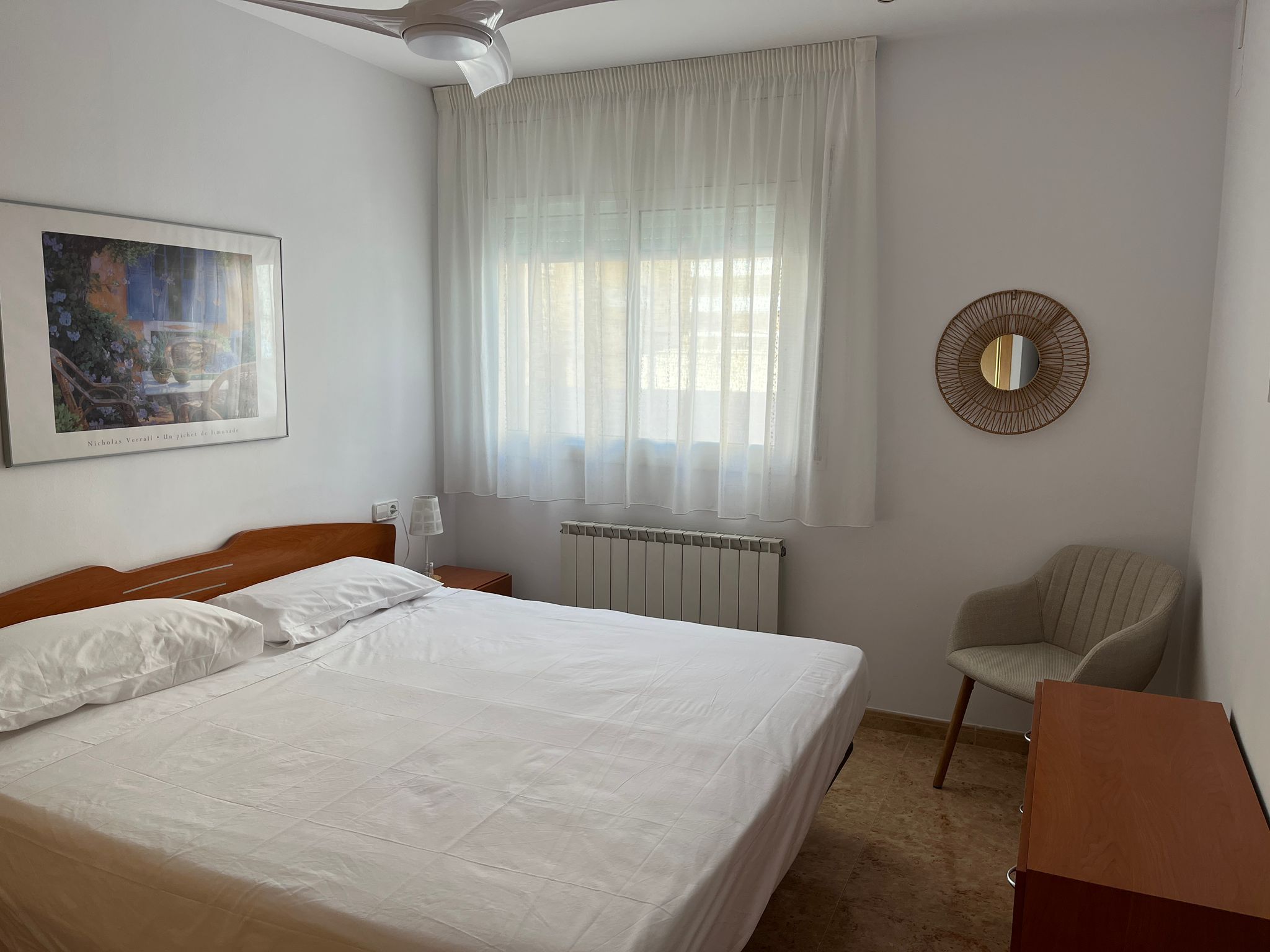 Una habitación de hotel con cama, una silla, un radiador, un ventilador de techo, una ventana con cortinas y un espejo montado en la pared.