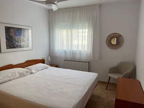 Quarto de hotel com cama, cadeira, radiador, ventoinha no teto, janela com cortinas e espelho fixado na parede.