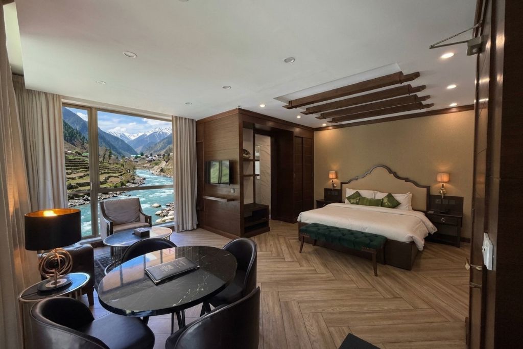 A hotel room with a bed, bedside tables, a desk with chairs, a view of a river and mountains, and a large window.