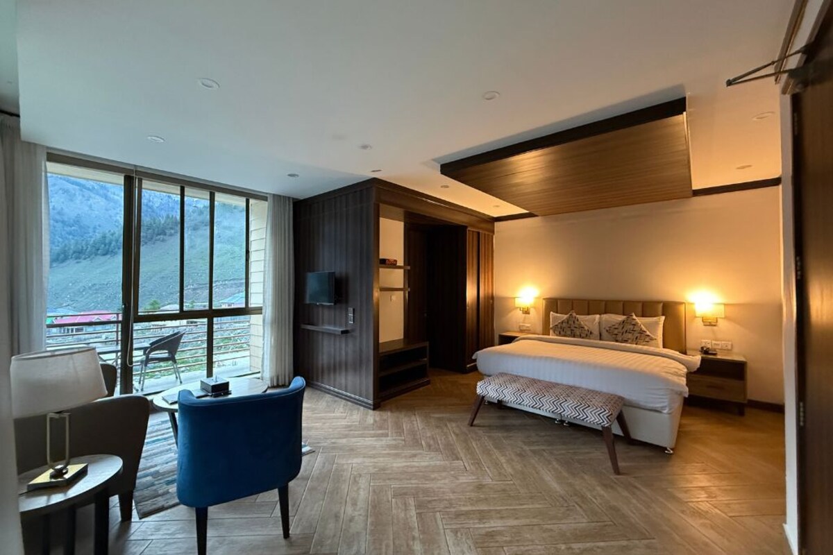 A modern hotel room with a large bed, a desk, and a chair. There is a TV mounted on the wall, and the room has a large window with a view of the outdoors.