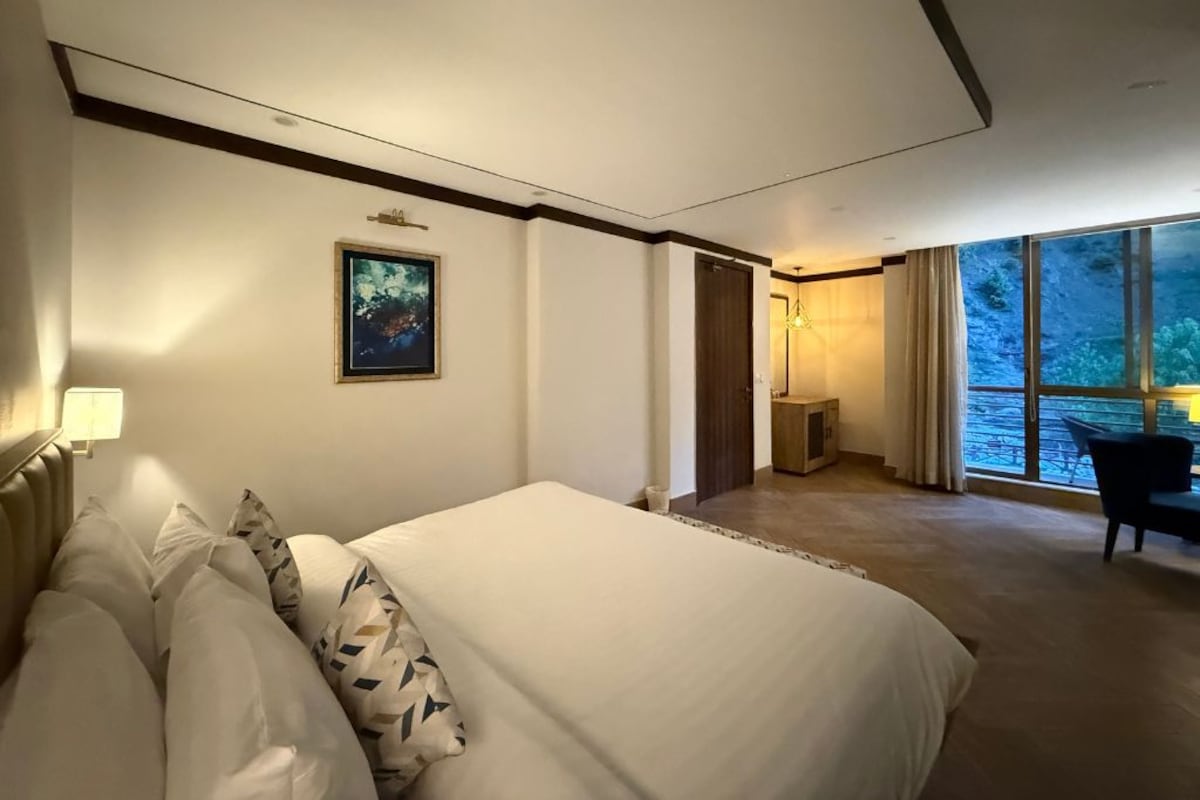 A hotel room with a large bed, a desk, a chair, and a view of the outdoors through a large window.