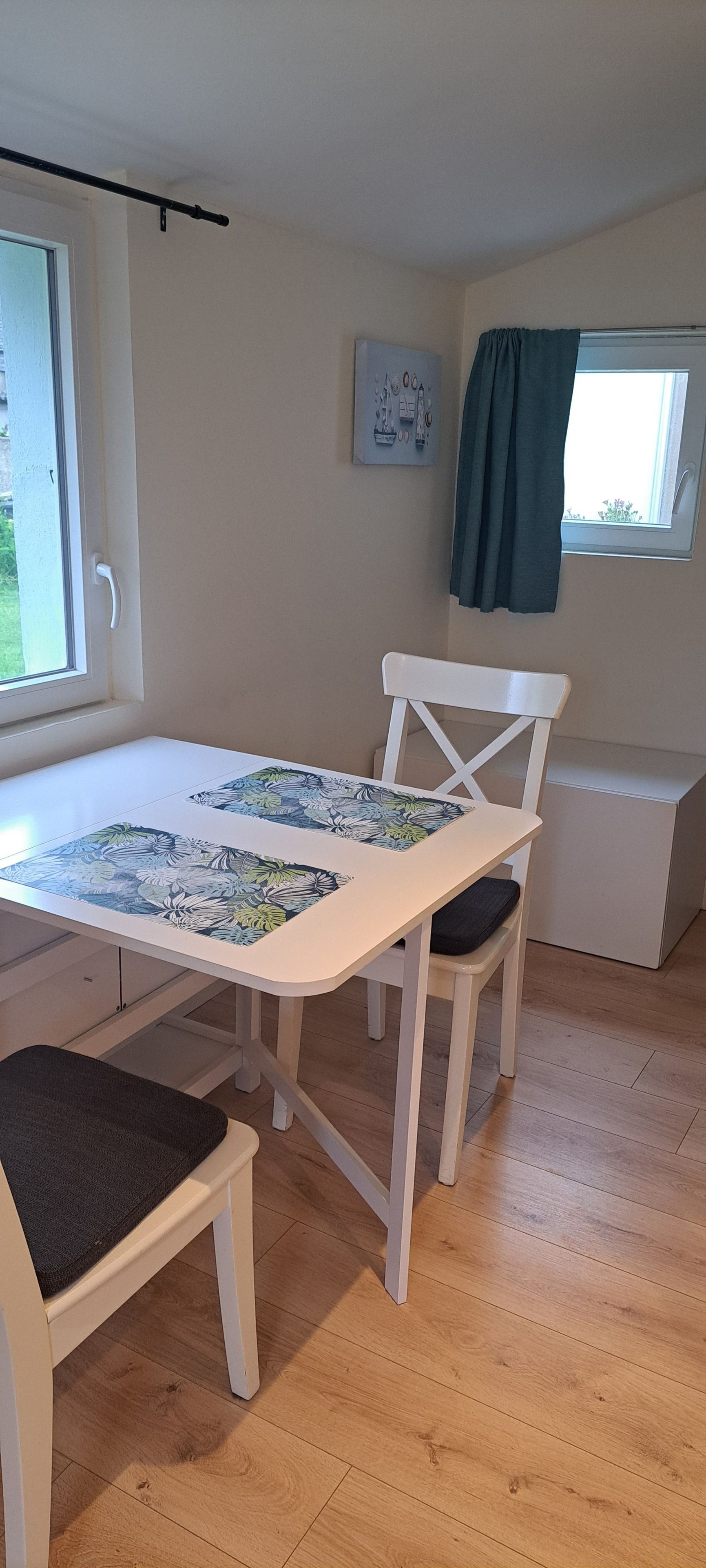 Una habitación con una mesa y sillas blancas, una ventana con cortina y una obra de arte colgada en la pared.