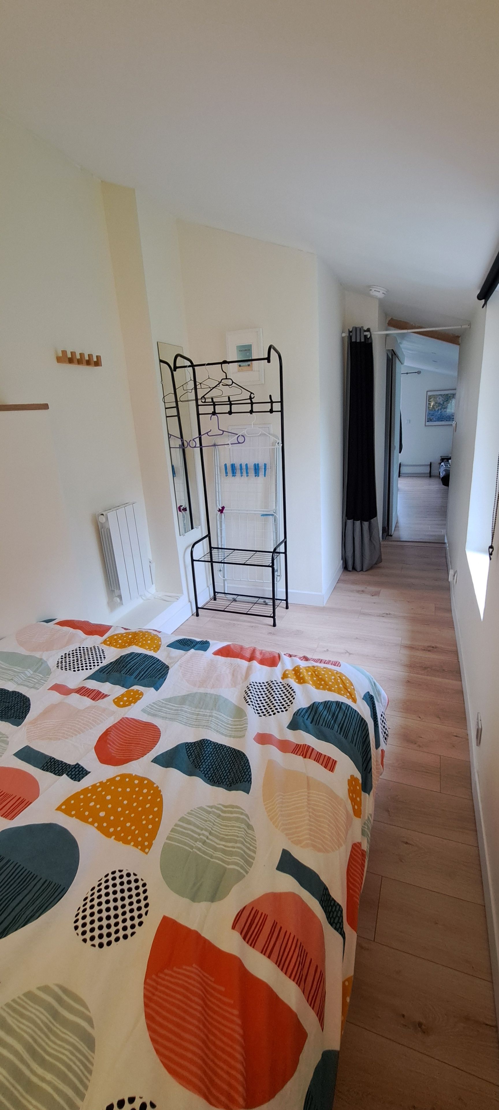 Et soveværelse med seng, et metalstativ til tøj, en radiator og en gang der fører til et andet rum.