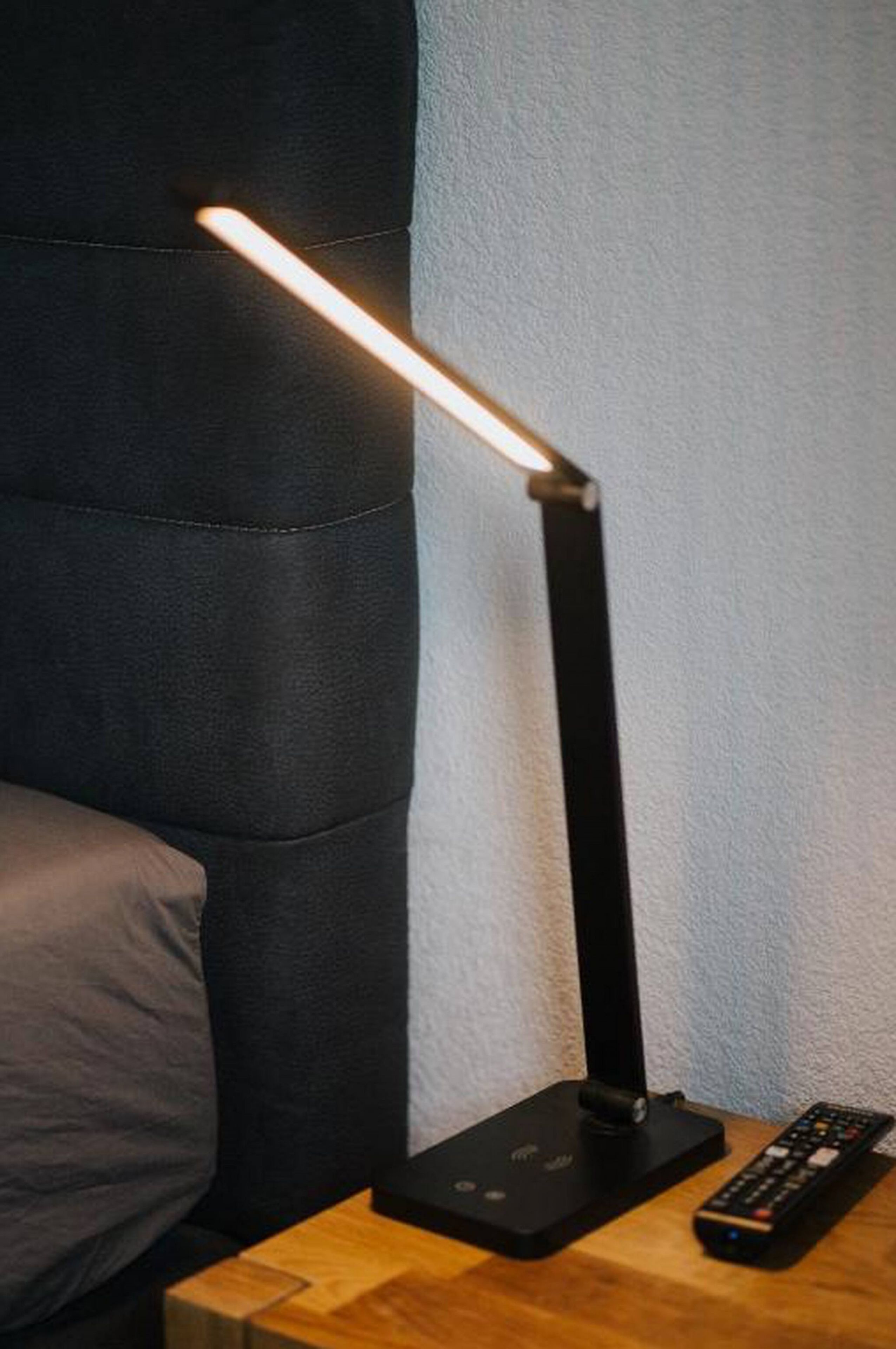 A black desk lamp with a rectangular base and a long, straight neck, positioned on a wooden surface next to a remote control.