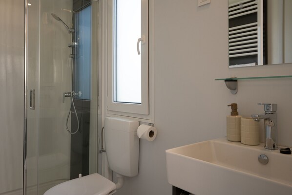 A bathroom with a glass shower enclosure, a toilet, a sink with a mirror above it, and a window. - Sonnendeck Tiny House Klagenfurt by WÖRTHWHILE (Klagenfurt am Wörthersee)