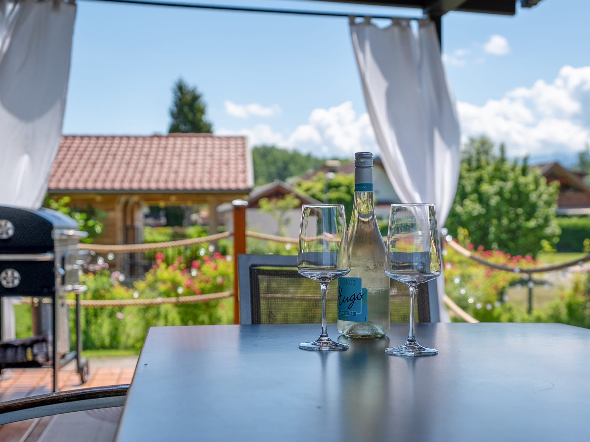A table with a bottle and two glasses, a grill, and a view of a house and garden.