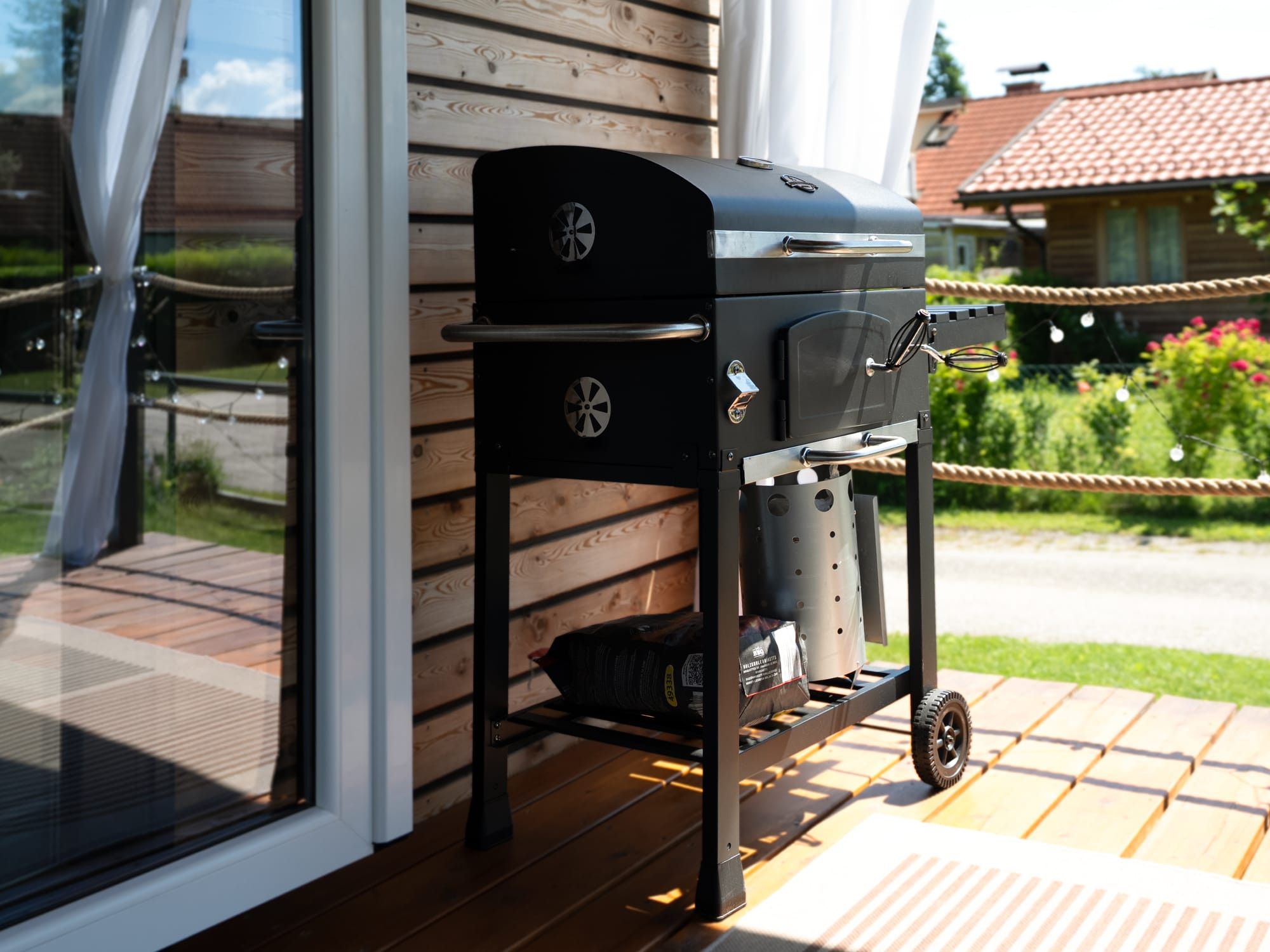 Ein Grill mit integriertem Stauraum und Rädern, platziert auf einer Holzterrasse neben einem Haus.
