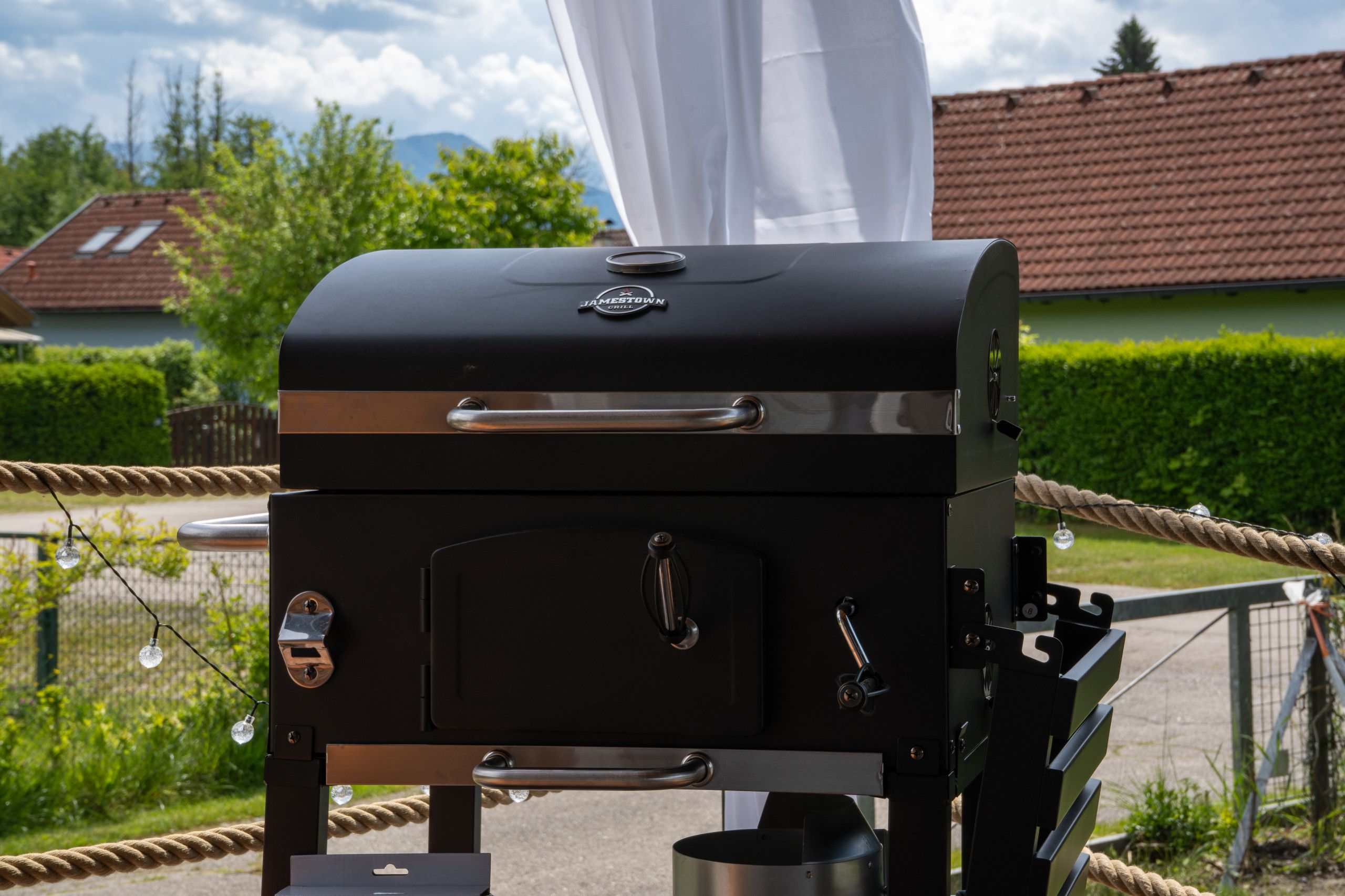 Un barbecue noir pour l’extérieur, doté d’une poignée et d’un support en métal pour les ustensiles, entouré d’une barrière faite de cordes.