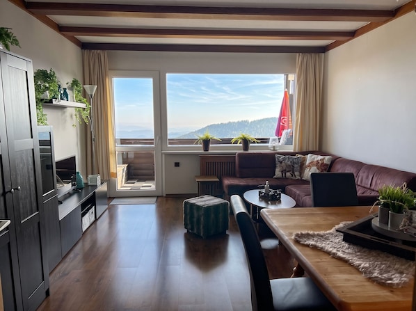 Una sala moderna con piso de madera, un sofá, una mesa de comedor y vistas a una cadena montañosa boscosa a través de amplias ventanas.