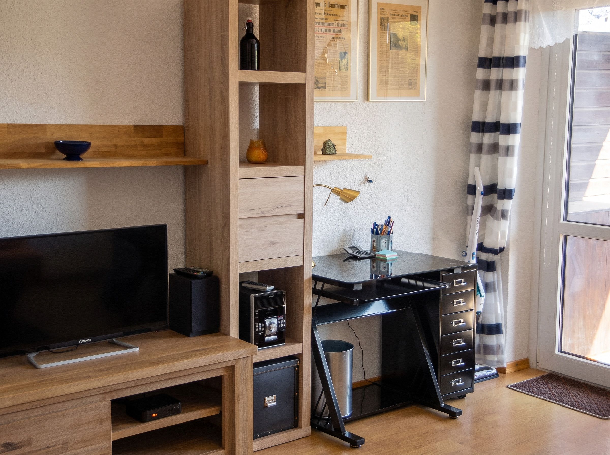 Un salon moderne doté d’un téléviseur à écran plat, d’étagères en bois, d’un bureau avec une chaise et d’une grande fenêtre avec des rideaux.