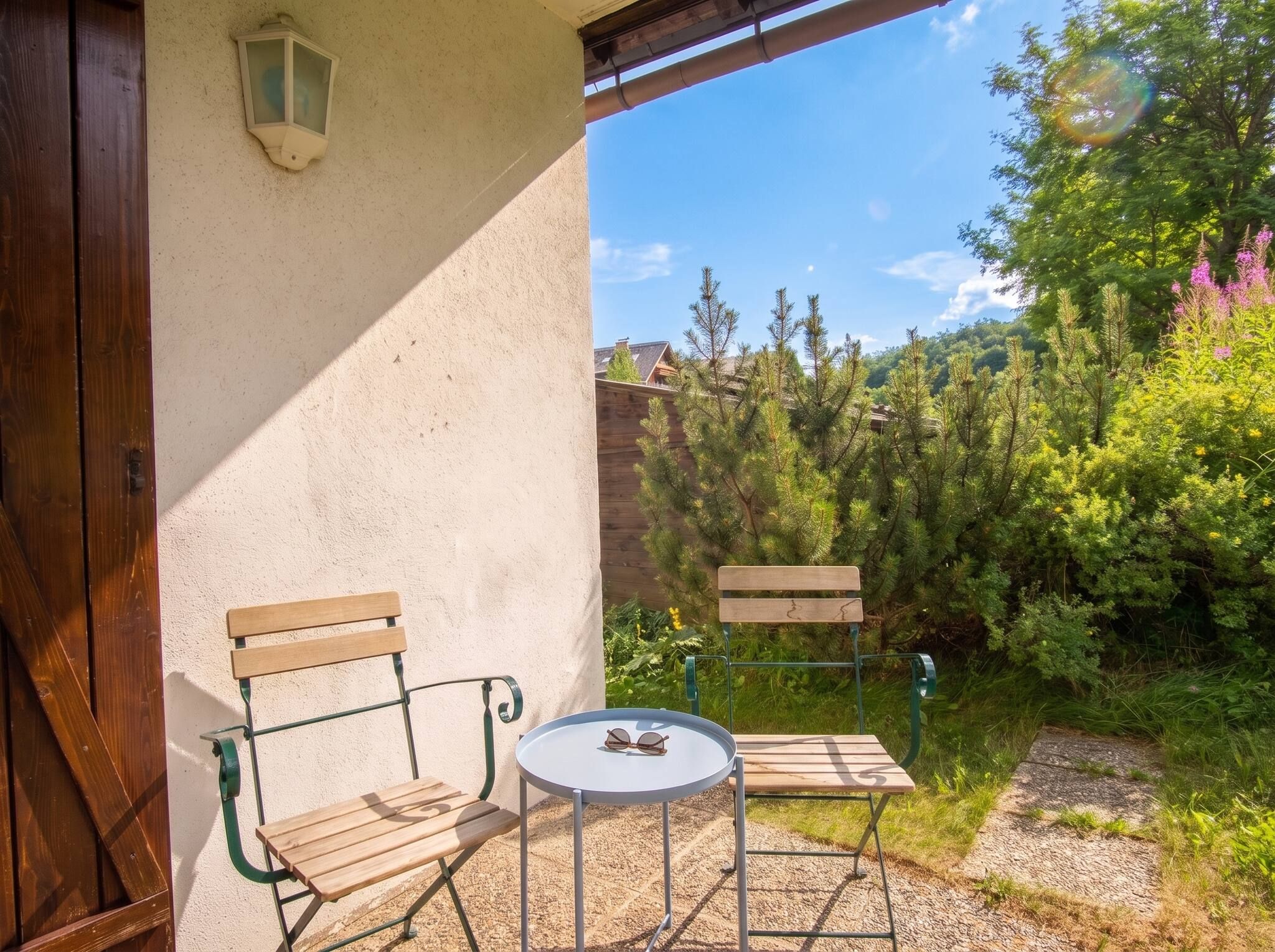 Un espace extérieur aménagé avec deux chaises en bois et une petite table ronde, une porte en bois et un luminaire fixé au mur.