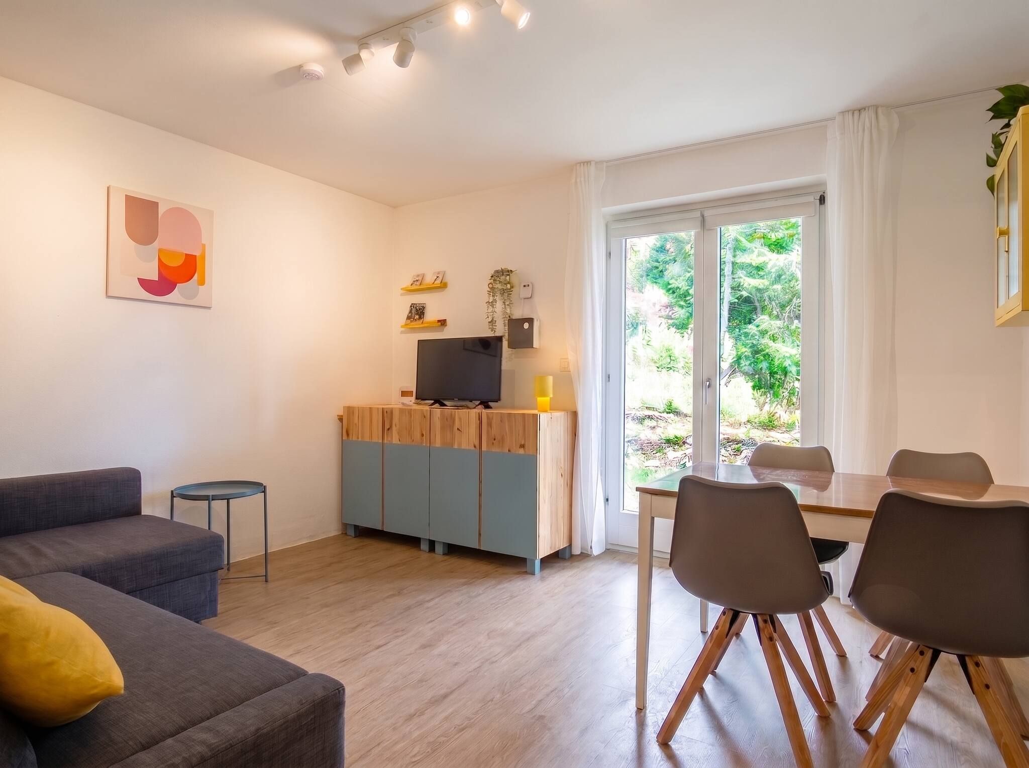 A modern living room with a sofa, a dining table with chairs, a TV on a wooden cabinet, and a large window with a view of greenery.
