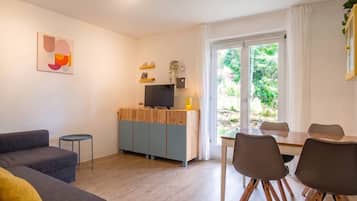A modern living room with a sofa, a dining table with chairs, a TV on a wooden cabinet, and a large window with a view of greenery.