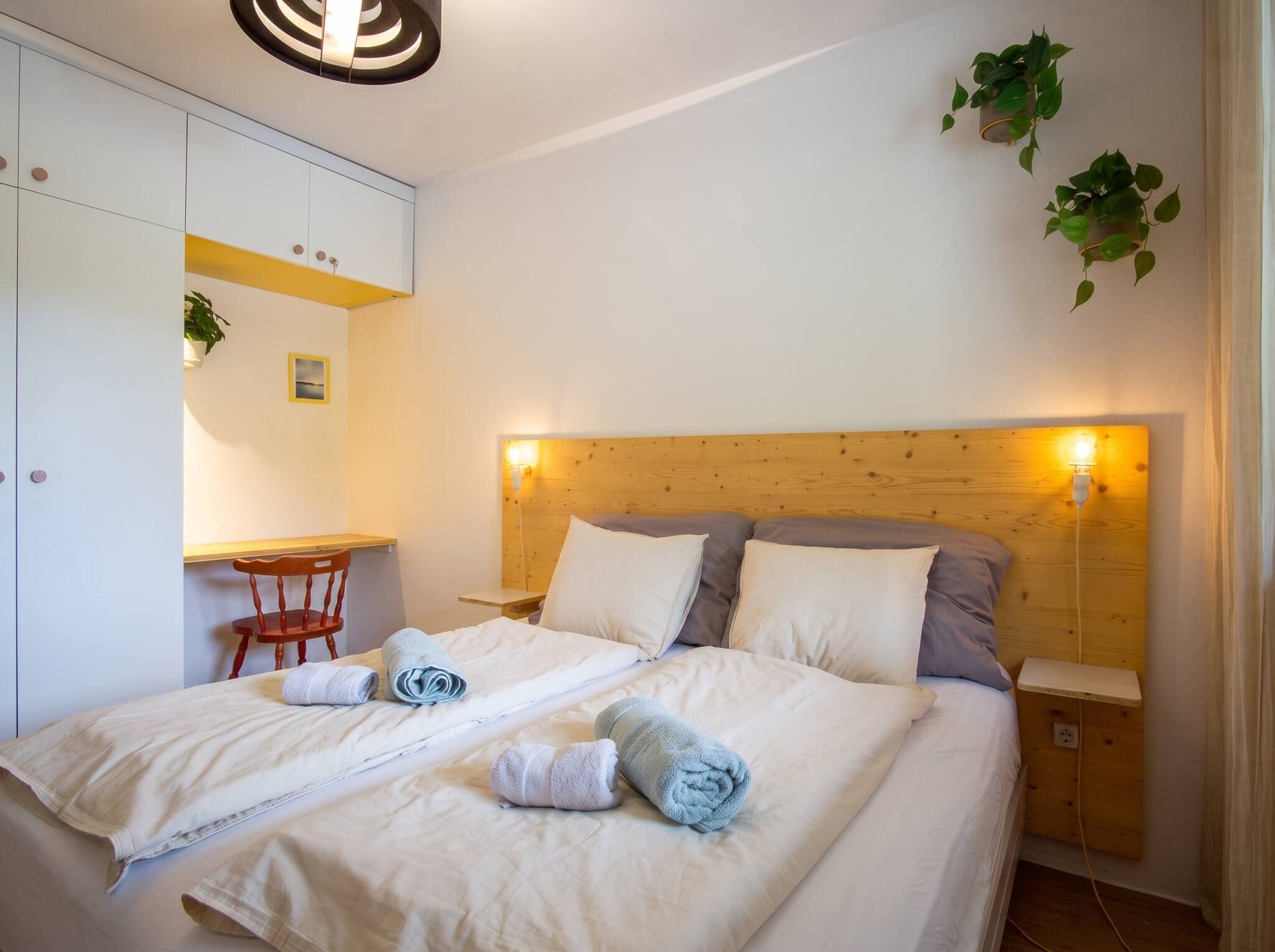 A neatly made bed with pillows and towels, a wooden headboard, a small table with a chair, and a wall-mounted plant.