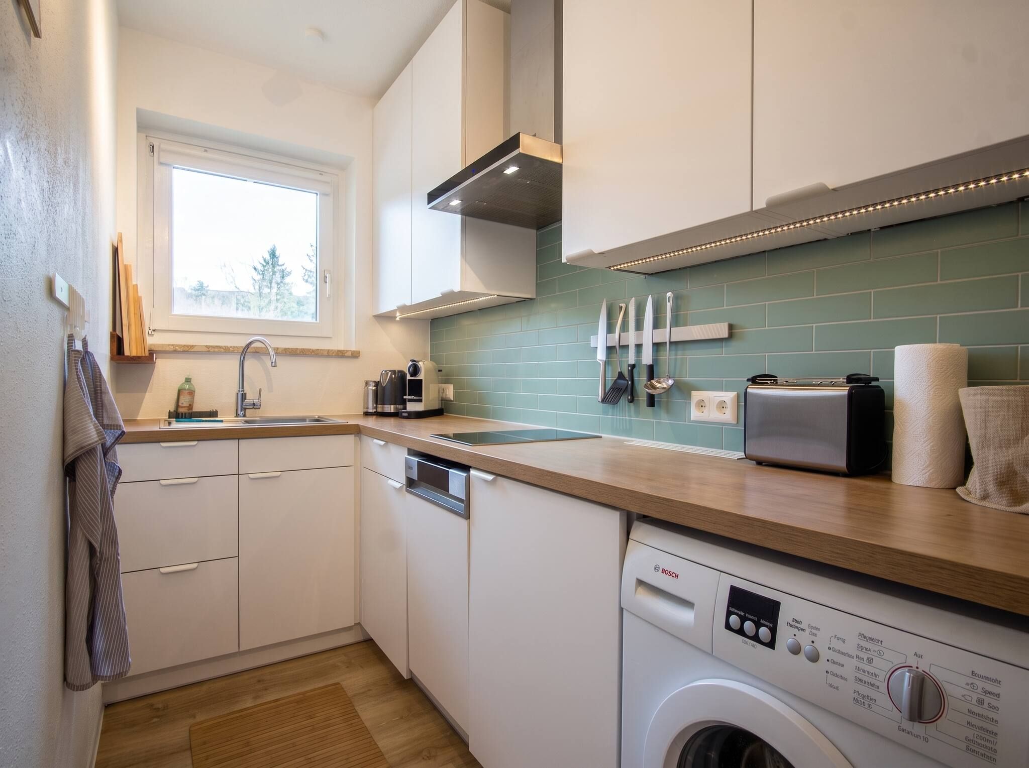 A modern kitchen with a dishwasher, a Bosch washing machine, and a window providing natural light.