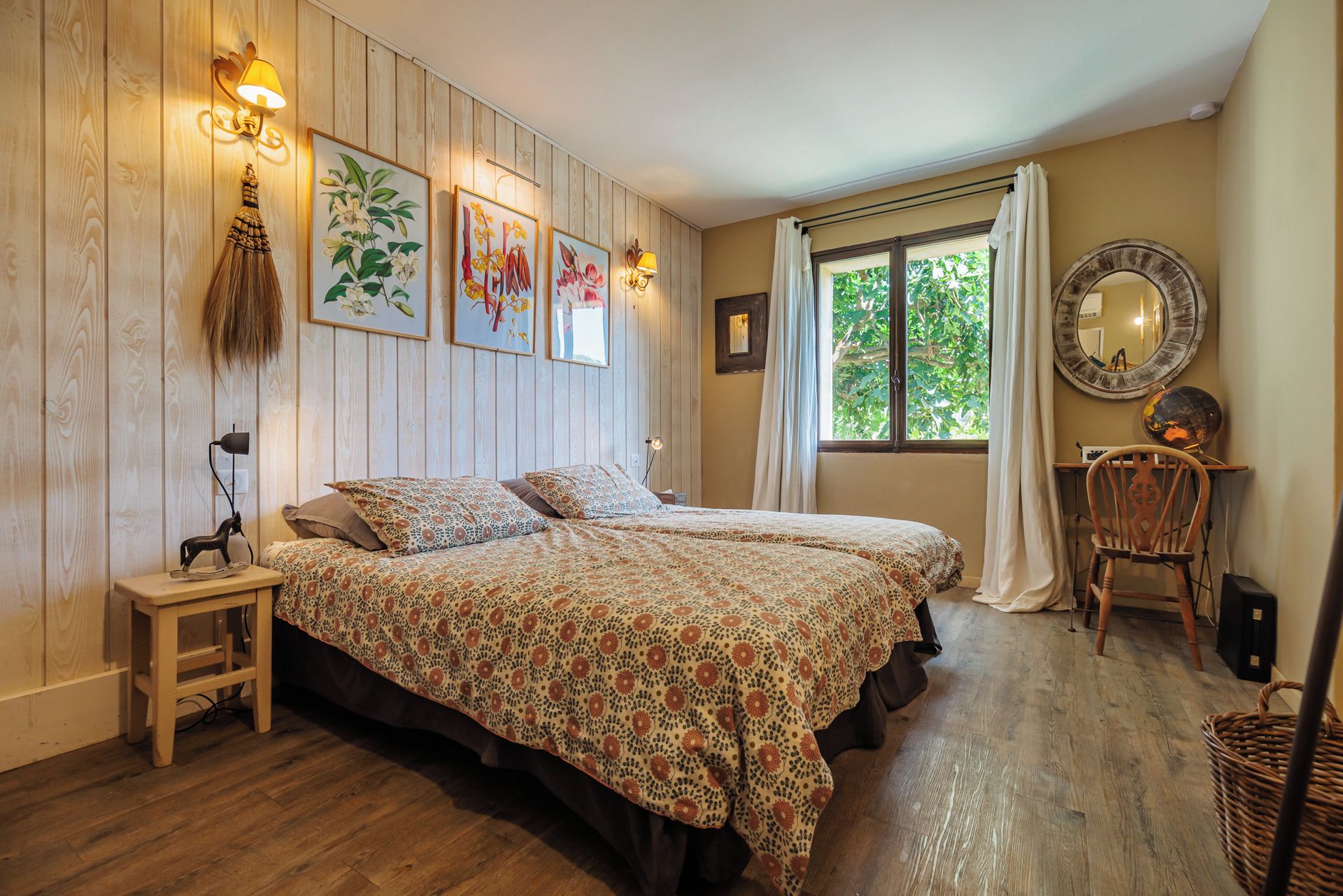 A bedroom with a wooden floor, a bed with floral bedding, a nightstand, a chair, a window with curtains, and wall-mounted lights.