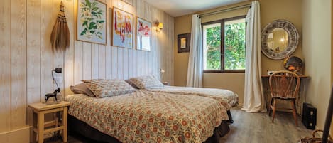 A bedroom with a wooden floor, a bed with floral bedding, a nightstand, a chair, a window with curtains, and wall-mounted lights.