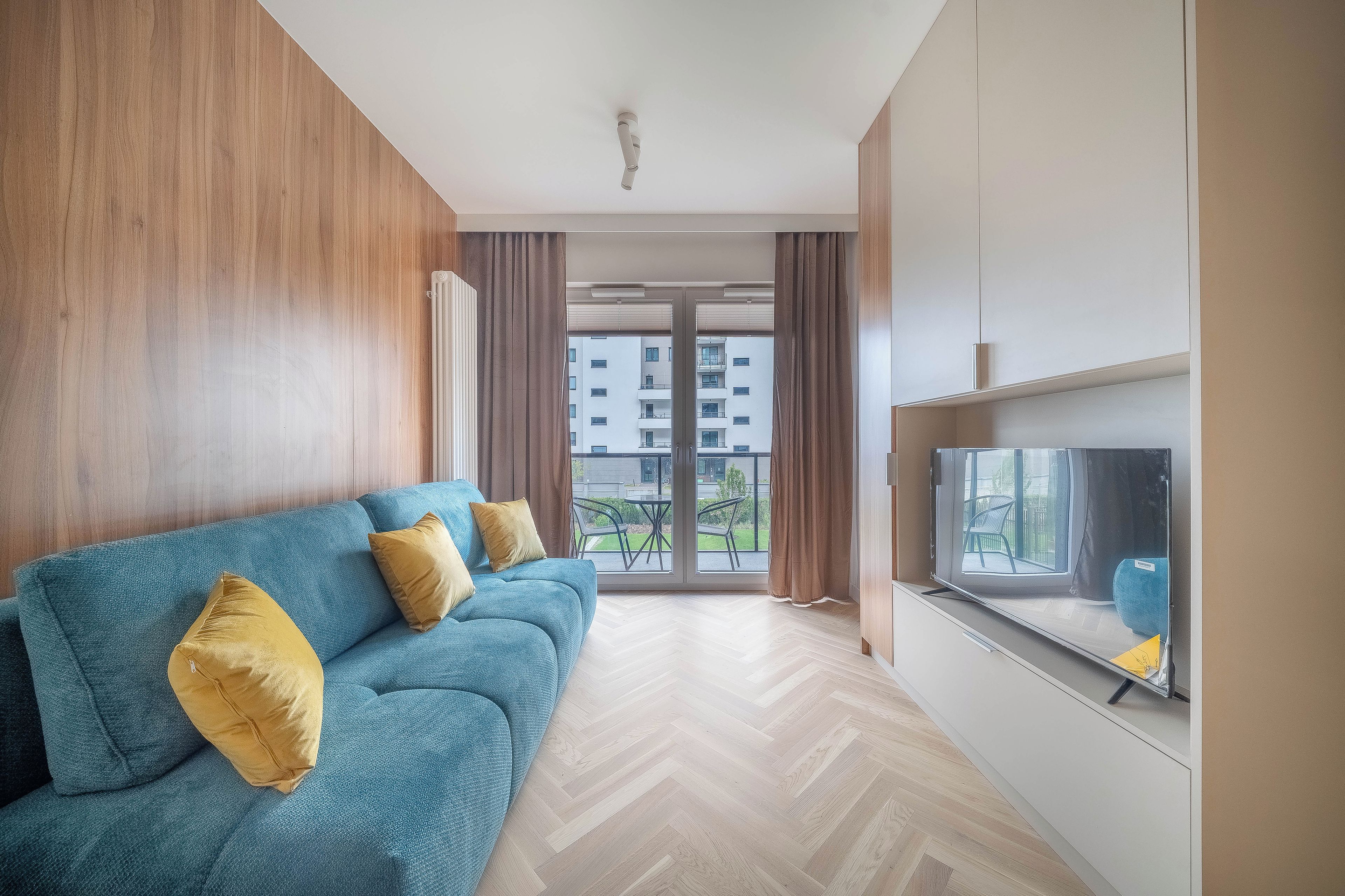 Uma sala de estar moderna com um sofá azul, televisão e vista para um prédio residencial.