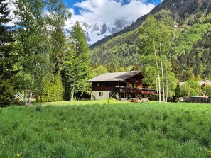 Eine Holzhütte mit Balkon, umgeben von üppigem Grün und Bergen im Hintergrund.