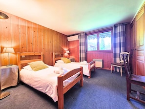 Ein Hotelzimmer mit zwei Holzbetten, einem kleinen Tisch, einem Sessel und einem Fenster mit Vorhängen.