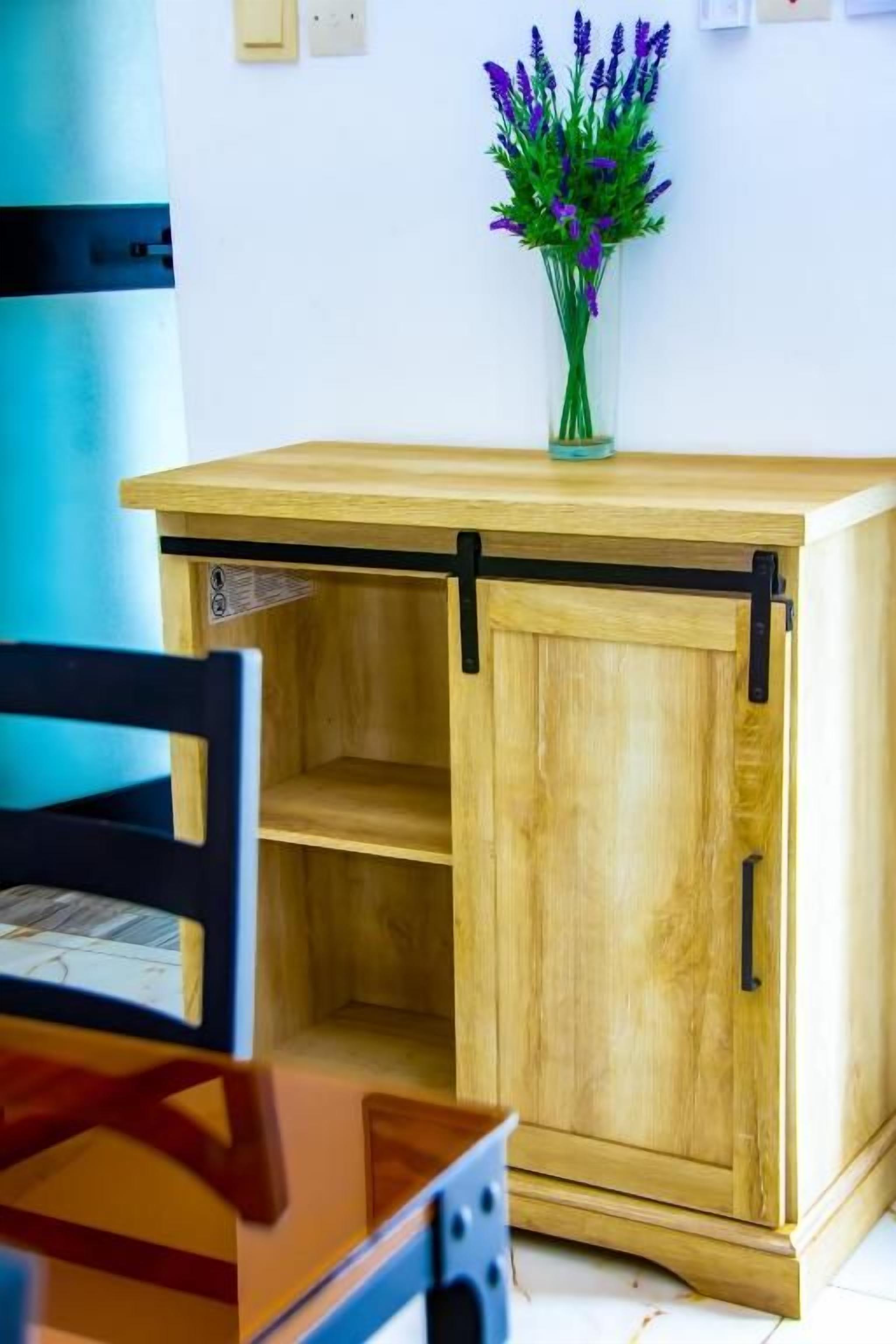 A wooden cabinet with a sliding door, a vase with purple flowers on top, and a blue cylindrical object to the left.