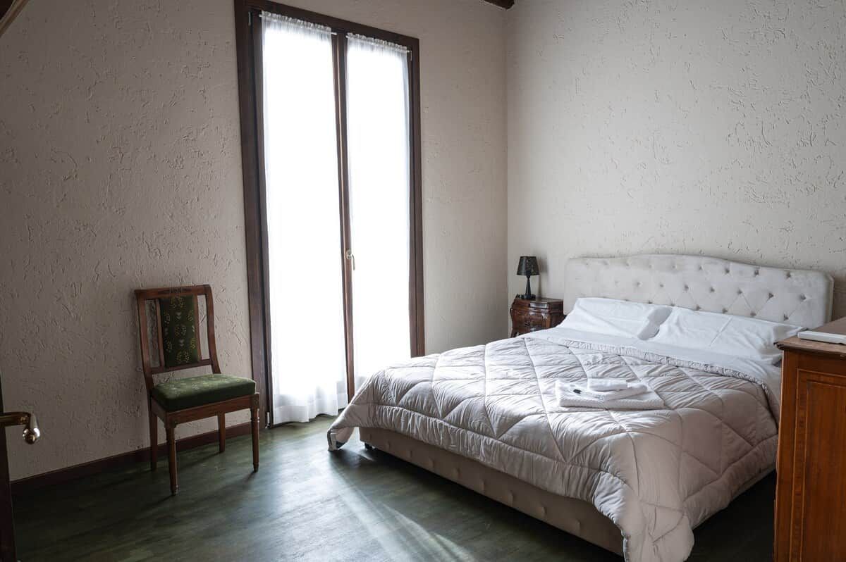 Un dormitorio con cama, una silla, una mesita de noche y una ventana.