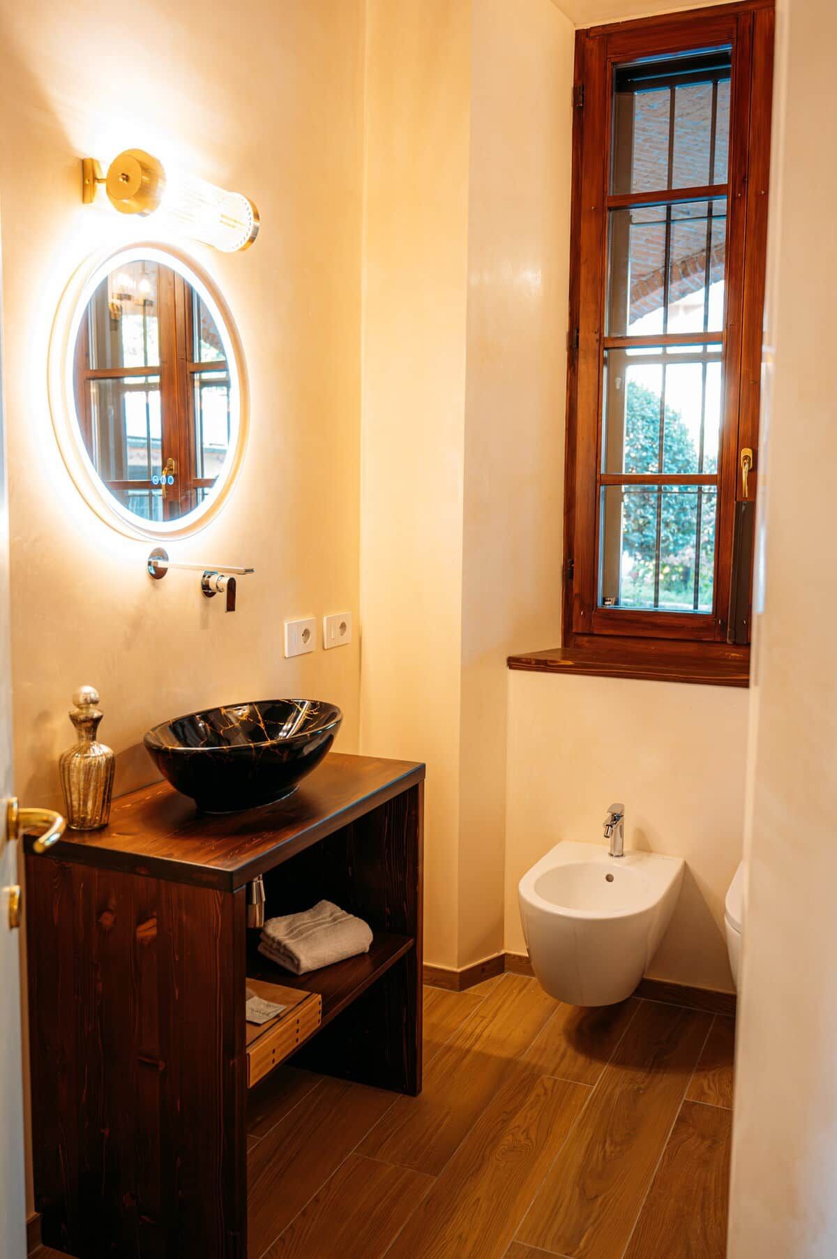 A bathroom with a wooden vanity, a round mirror, a bidet, and a window with a view.