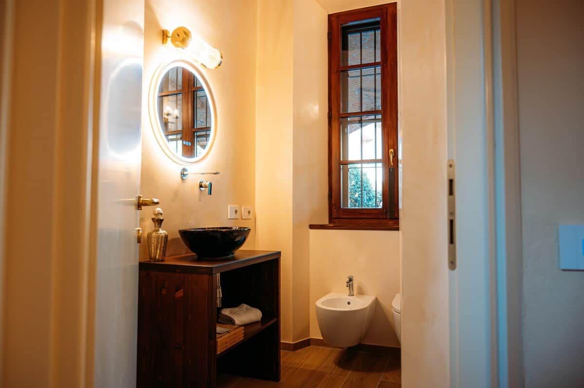 Une salle de bain avec un meuble-lavabo en bois, un miroir rond, un bidet et une fenêtre donnant sur la verdure.