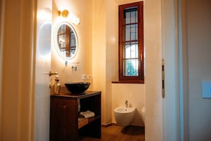 A bathroom with a wooden vanity, a round mirror, a bidet, and a window with a view of trees. - Love's Nest | A Romantic Getaway on Lake Maggiore (Ispra)