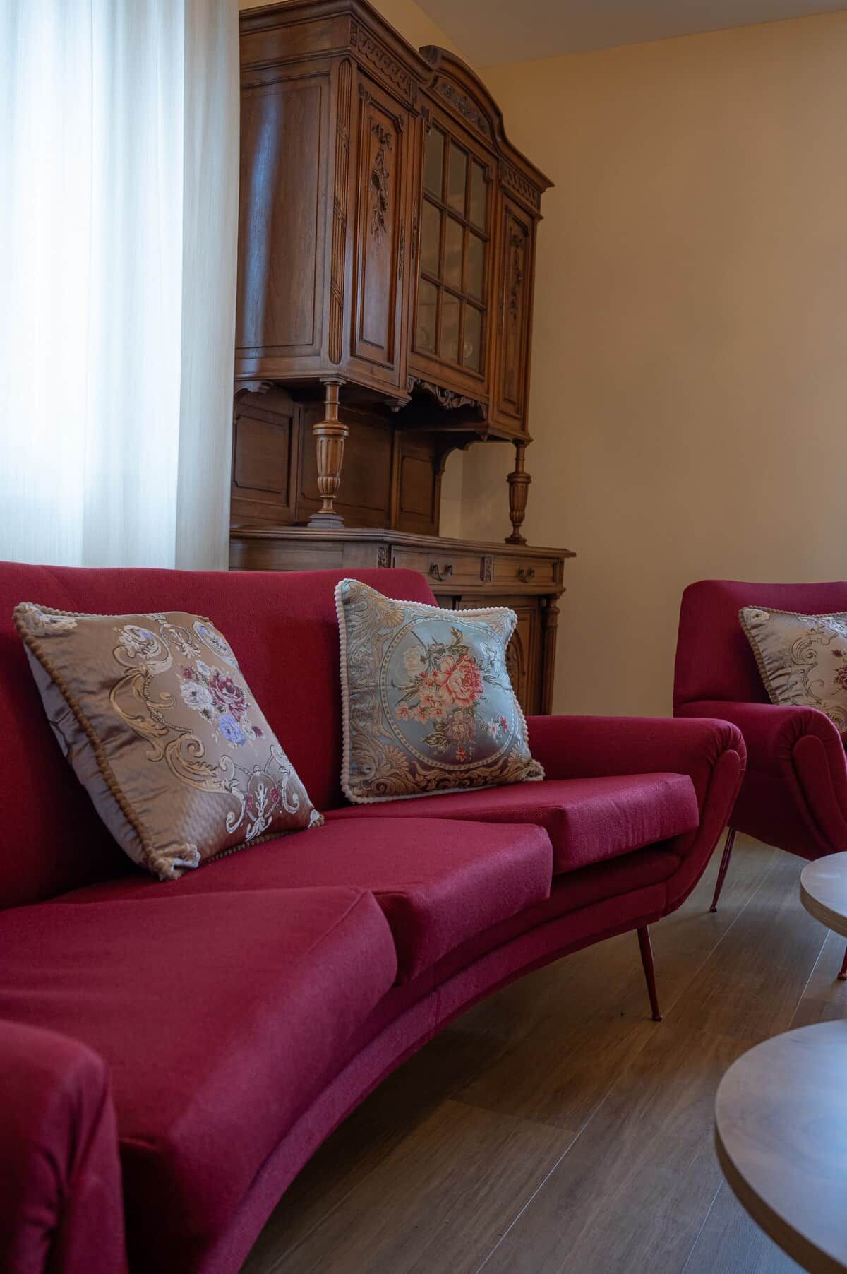 Um sofá vermelho com almofadas florais, um armário de madeira e uma mesa redonda em um quarto com piso de madeira.