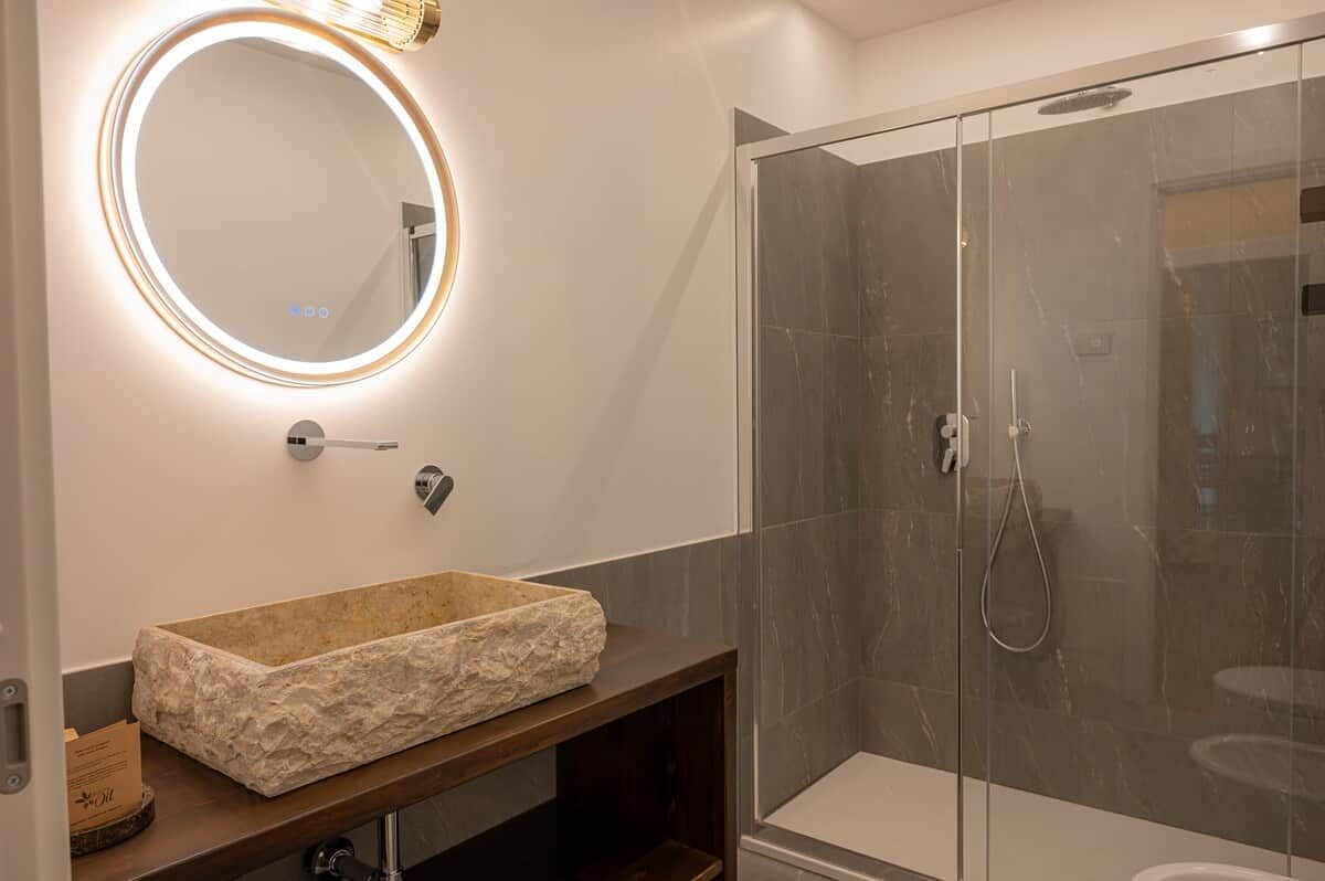 Une salle de bain avec un lavabo en pierre, un miroir rond avec éclairage intégré et une douche à parois de verre.